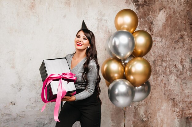 Un ritratto a figura intera di una ragazza di compleanno con i capelli ricci in scarpe col tacco alto che tiene un regalo in una scatola Elegante giovane donna tiene un regalo per una vacanza palloncini