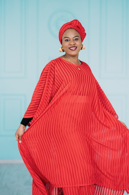 Un ritratto di bella signora africana che indossa abiti eleganti rossi e copricapo, in posa con un sorriso felice su sfondo azzurro dello studio.