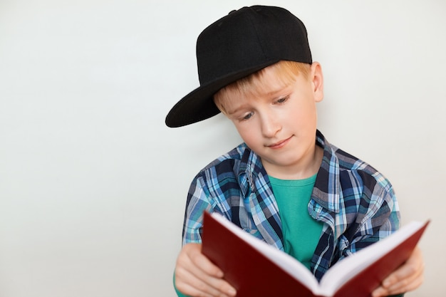 Un ritratto di curioso ragazzino in berretto nero e controllato camicia colorata lettura enciclopedia guardando nel libro con interesse