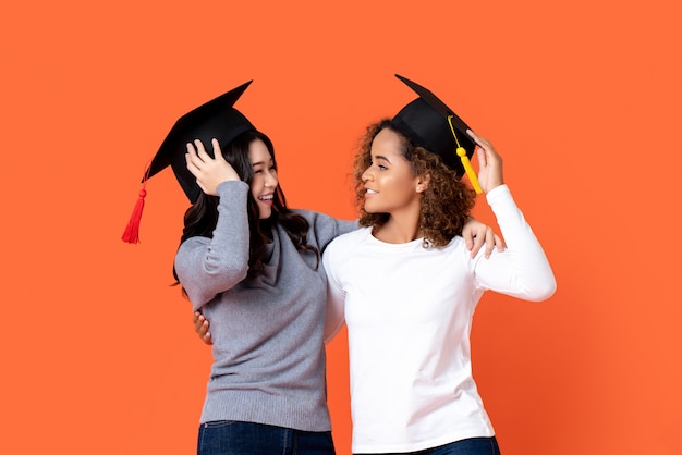 Un ritratto di due donne razza mista felici che si laureano tenendo la protezione di graduazione che se lo esamina in parete isolata arancia