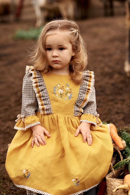 Un ritratto di un bambino carino in un bel vestito giallo seduto su una panchina in una fattoria su uno sfondo di capre