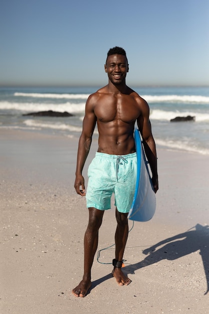 Un ritratto di un uomo afroamericano felice e attraente che si gode il tempo libero sulla spiaggia in una giornata di sole, sorridendo, divertendosi, in piedi con la sua tavola da surf, il sole che splende su di lui.