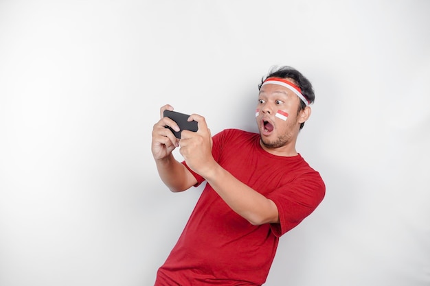 Un ritratto di un uomo asiatico scioccato che indossa una fascia per la testa che tiene in mano il suo telefono mentre la sua bocca è spalancata isolata da uno sfondo bianco