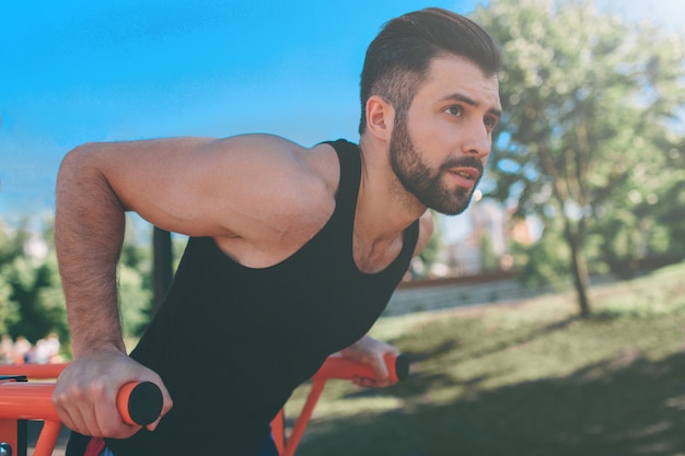 Un ritratto di un uomo muscoloso con la barba concentrato in abiti neri allenamento facendo immersioni su barre parallele. Equipaggia la forma fisica con il cielo blu sullo sfondo e lo spazio aperto intorno a lui. Sport e crossfit.