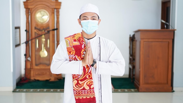 Un ritratto di un uomo musulmano asiatico si alza in una posa di saluto alle mani di Namaste, accogliendo gli ospiti, Ied Fitr saluta alla moschea. Indossare la maschera