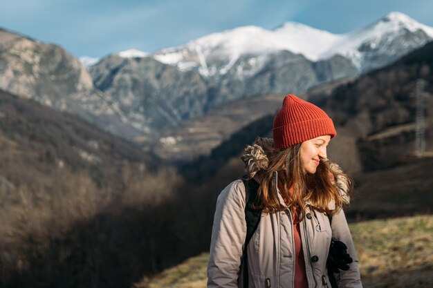 Un ritratto di un viaggiatore sorridente sullo sfondo delle montagne innevate donna felice in berretto rosso che si scalda e si gode la luce del sole durante le escursioni in montagna