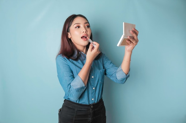 Un ritratto di una bella giovane donna asiatica che applica il rossetto sulle labbra isolate da uno sfondo blu