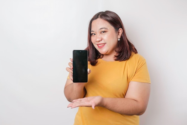 Un ritratto di una donna asiatica di grandi dimensioni felice sorride e mostra lo spazio della copia sul suo smartphone che indossa una camicia gialla isolata da uno sfondo bianco