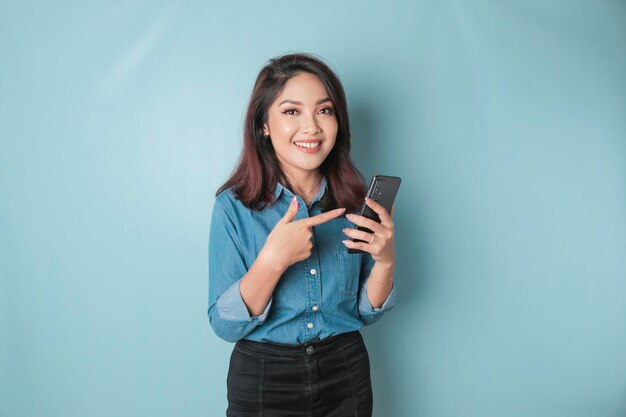 Un ritratto di una donna asiatica felice sorride e tiene in mano il suo smartphone indossando una maglietta blu isolata da uno sfondo blu