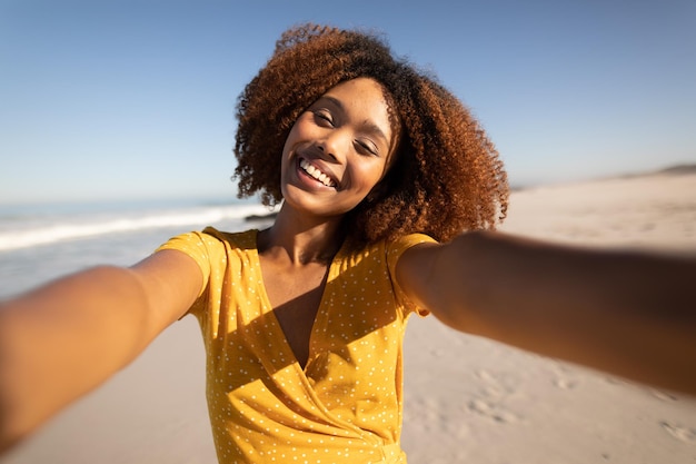 Un ritratto di una donna di razza mista felice e attraente che si gode il tempo libero sulla spiaggia in una giornata di sole, indossa un abito giallo, in piedi sulla sabbia, prendendo selfie con il sole che splende sul viso.
