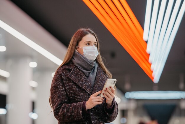 Un ritratto ravvicinato di una donna con una maschera medica è seduto alla stazione della metropolitana con uno smartphone e in attesa di un treno. Una ragazza con una mascherina chirurgica sta mantenendo le distanze sociali in metropolitana.