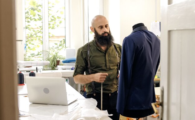 Un sarto in un laboratorio di cucito al lavoro. Il suo studio è soleggiato. Tessuti colorati, capi di abbigliamento sono visibili.