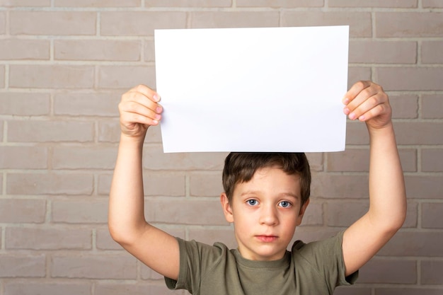 Un serio ragazzo europeo di 5 anni tiene un poster vuoto sopra la sua testa Sfondo in mattoni beige Il concetto di protesta Layout