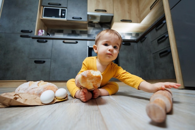 Un simpatico bambino di un anno è seduto in cucina e mangia un pane lungo o una baguette in cucina Il primo pasto di pane da parte di un bambino Il pane fa bene ai bambini