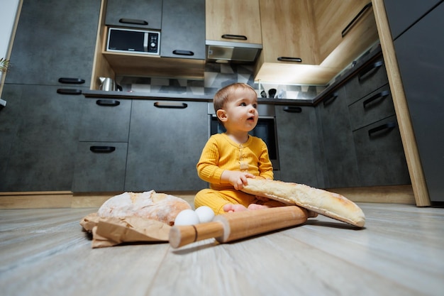 Un simpatico bambino di un anno è seduto in cucina e mangia un pane lungo o una baguette in cucina Il primo pasto di pane da parte di un bambino Il pane fa bene ai bambini