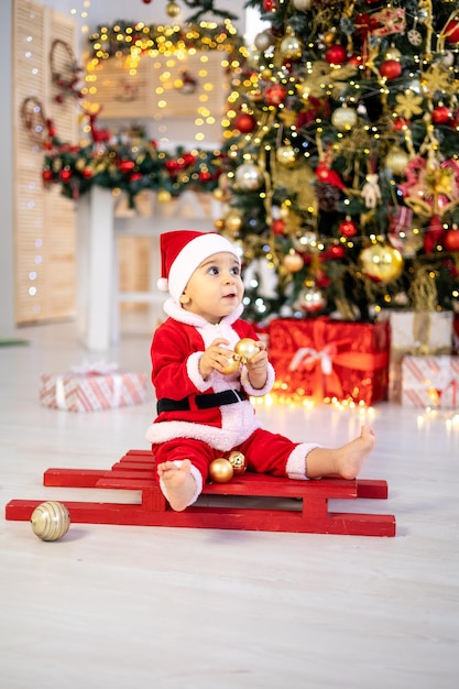 Un simpatico bambino in costume da Babbo Natale è seduto su una slitta sotto un albero di Natale festivo con regali nel soggiorno di casa un bambino felice festeggia il Natale e il nuovo anno a casa
