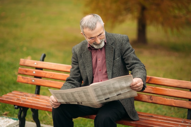 Un simpatico nonno con una bella barba e una giacca grigia si siede su una panchina del parco e legge a