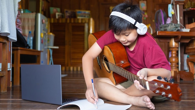 Un simpatico ragazzo asiatico che indossa le cuffie impara la musica e scrive canzoni usando una chitarra acustica a casa