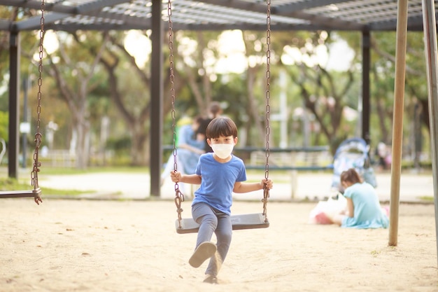 Un simpatico ragazzo asiatico che indossa una maschera sta giocando su un'altalena nel parco giochi durante il giorno in estate Attività all'aperto Il gioco fa credere alle idee Istruzione esterna ritratto