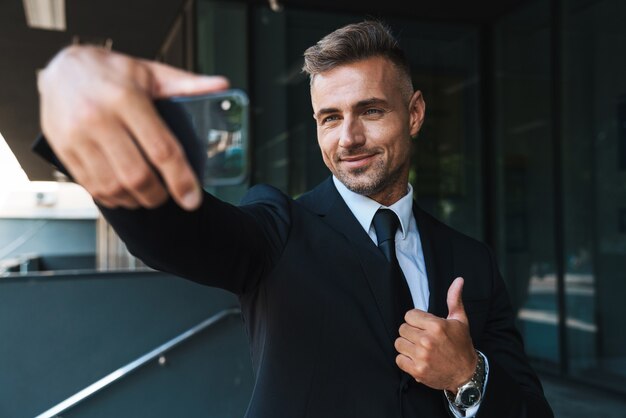 un sorridente ottimista maturo uomo d'affari dai capelli grigi all'aperto in strada vicino al centro business prendere selfie dal telefono cellulare che mostra i pollici in su gesto.