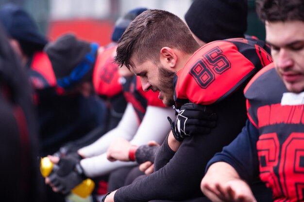 Un stanco giocatore di football americano che tiene la spalla infortunata mentre riposa con i compagni di squadra dopo un duro allenamento sul campo