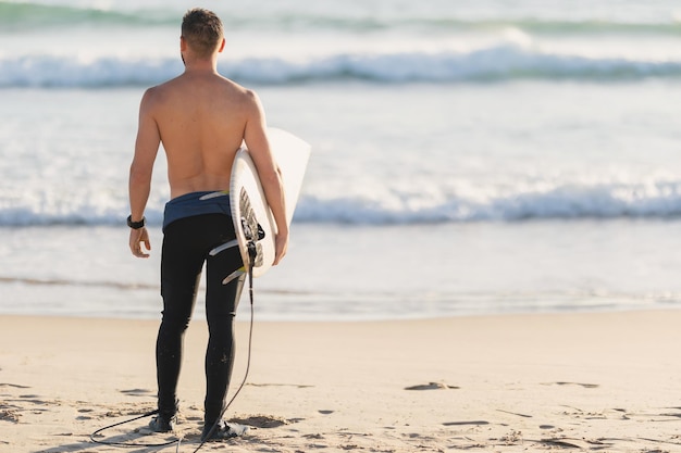 Un surfista atletico con il torso nudo in piedi vicino alla vista posteriore dell'oceano