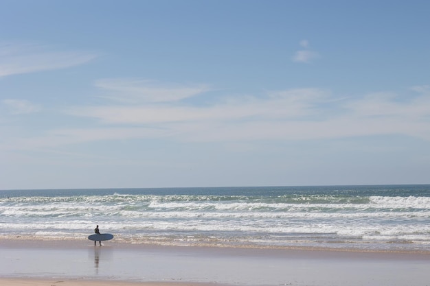 Un surfista solitario cammina lungo la spiaggia fino al mare con in mano una tavola