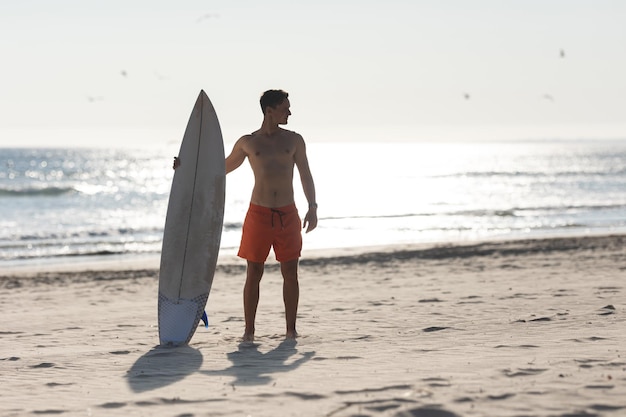 Un surfista uomo senza camicia in piedi sulla spiaggia che tiene una tavola da surf