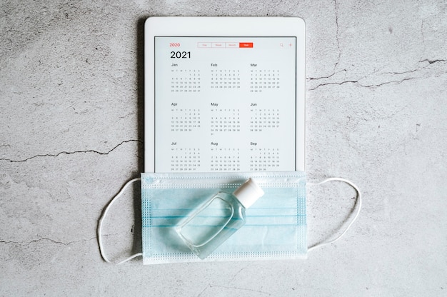 Un tablet con un calendario aperto per l'anno 2021 e maschera medica protettiva e disinfettante per le mani su un grigio