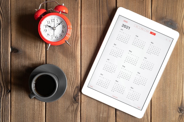 Un tablet con un calendario aperto per l'anno 2021, una tazza di caffè e una sveglia rossa su uno sfondo di tavola di assi di legno