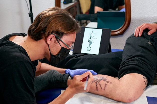 Un tatuatore che guarda un tablet e disegna un tatuaggio sulla gamba di un maschio in uno studio