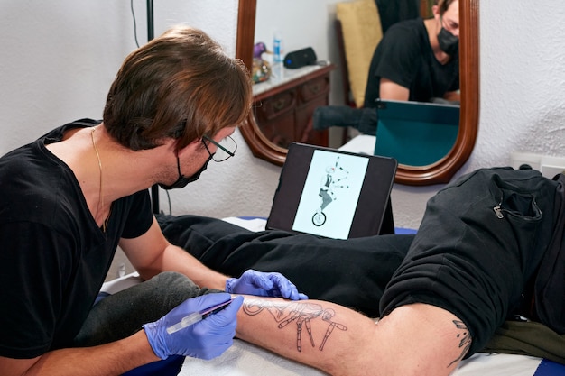 Un tatuatore in guanti blu che guarda un tablet e fa un tatuaggio sulla gamba di un maschio in uno studio