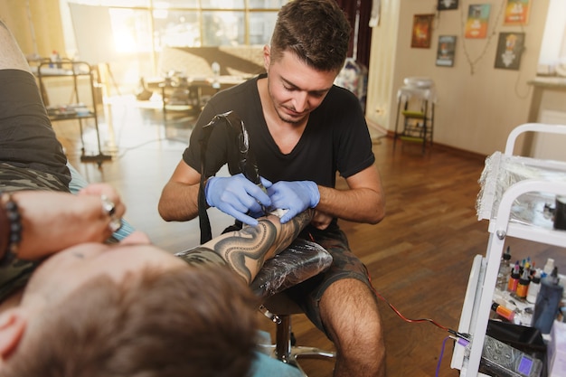 Un tatuatore professionista che fa un tatuaggio sul braccio di un giovane a macchina con inchiostro nero black