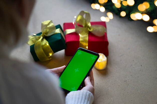 Un telefono con uno schermo verde nelle mani di una donna con un maglione bianco, una confezione regalo rossa e verde e luci bokeh sul tavolo. Avvicinamento. Concetto di shopping online per Natale e Capodanno