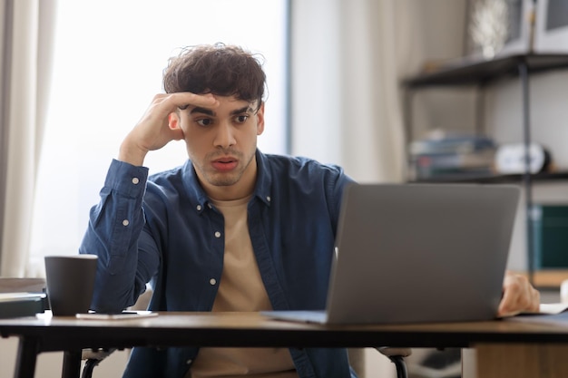 Un tizio del Medio Oriente annoiato seduto davanti al portatile che odia il lavoro in casa.
