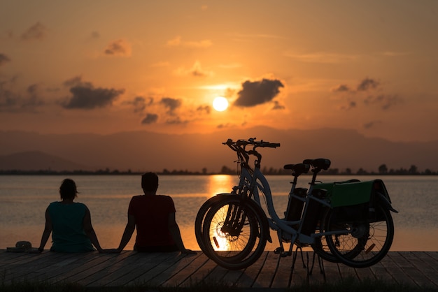 Un tramonto con una coppia stagliata seduta accanto alle loro biciclette in un molo in riva al mare
