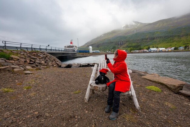 Un turista con un cappotto rosso si siede su una panchina e scatta foto del ponte Seydisfjrur Islanda