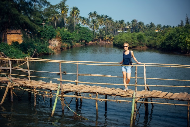 Un turista femminile in shorts in denim con taglio di capelli corto in giornata di sole tropicale. La giovane ragazza graziosa sta sul ponte di legno sopra il piccolo fiume contro la giungla e il cielo blu della palma.