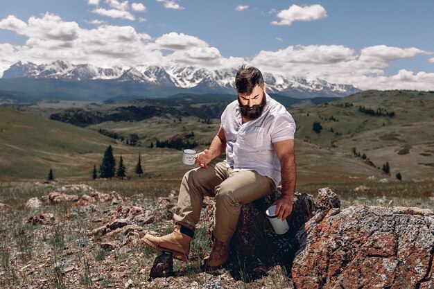 Un uomo adulto brutale con la barba in una camicia bianca tiene una tazza e un thermos per un'escursione seduto su una pietra con uno sguardo lanuginoso sullo sfondo di un paesaggio montano naturale.