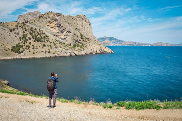 Un uomo adulto con attrezzatura fotografica si trova sul bordo di una scogliera Sotto c'è una pittoresca baia marina con barche da pesca