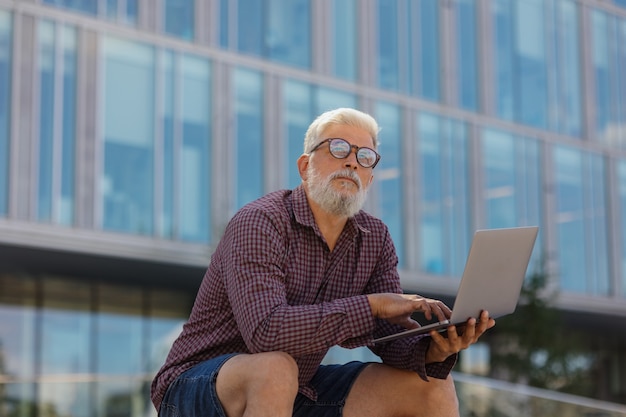 Un uomo adulto dai capelli grigi con la barba è seduto in ufficio e lavora su un laptop un anziano d'affari