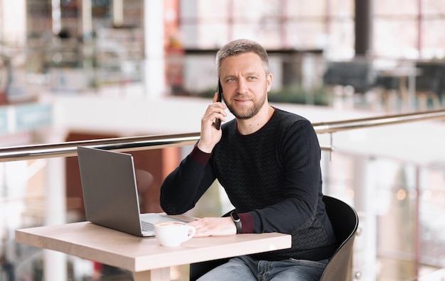 Un uomo adulto di successo lavora su un laptop e parla al telefono in una caffetteria