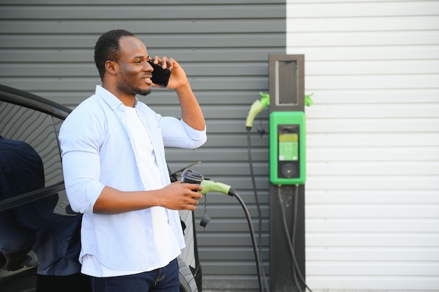 Un uomo africano è in piedi vicino a un'auto elettrica in attesa che venga caricata in una stazione di ricarica e usa il telefono