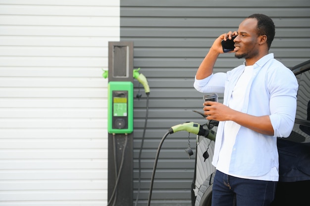 Un uomo africano è in piedi vicino a un'auto elettrica in attesa che venga caricata in una stazione di ricarica e usa il telefono