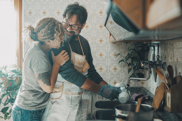 Un uomo aiuta la moglie nelle faccende domestiche a lavare i piatti in cucina La donna apprezza l'aiuto del marito nelle attività ricreative delle pulizie Coppia sorride e abbraccia con amore dentro la casa pulendo insieme