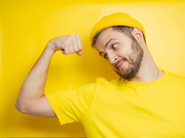 Un uomo allegro alza il braccio e mostra i suoi muscoli, sorride felicemente, dimostra la sua forza isolata su uno sfondo giallo. Un giovane energico