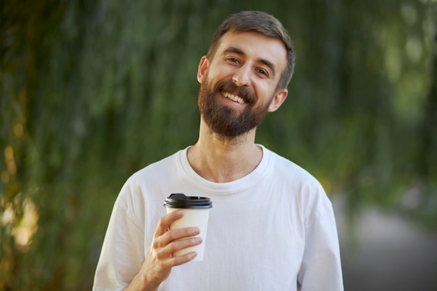 Un uomo allegro con una maglietta bianca beve caffè da una tazza di carta bianca. Una tazza di carta bianca