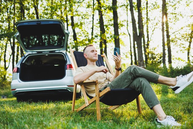 Un uomo allegro in occhiali si siede su una sedia in natura e lavora in vacanza lavoro online su un laptop portatile Lavoro freelance