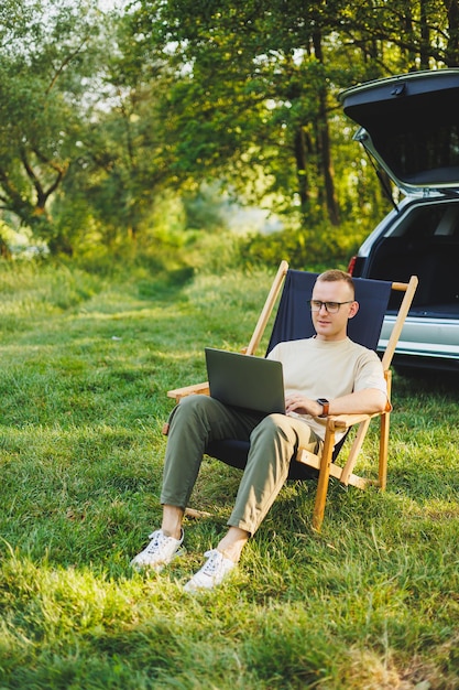 Un uomo allegro si siede su una sedia nella natura e lavora online su un laptop Un uomo viaggia e lavora a distanza su un computer portatile Lavoro d'ufficio nella natura