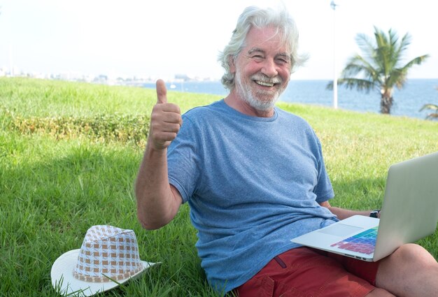 Un uomo anziano attraente e sorridente che lavora all'aperto con un laptop seduto in un prato verde